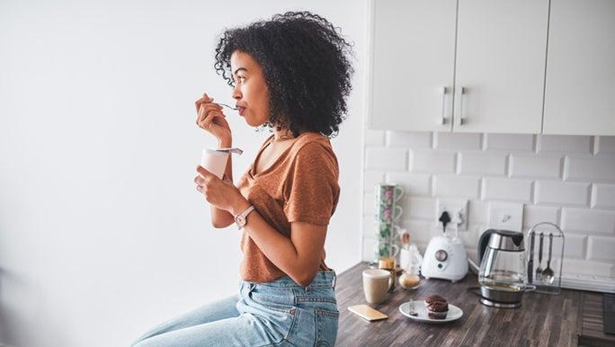 A woman with a dark complexion is sitting on a kitchen bench eating yoghurt which may contain probiotics for gut health